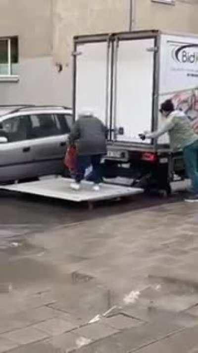 Guys use a lift gate on a truck to help a senior get into her car without getting her feet wet