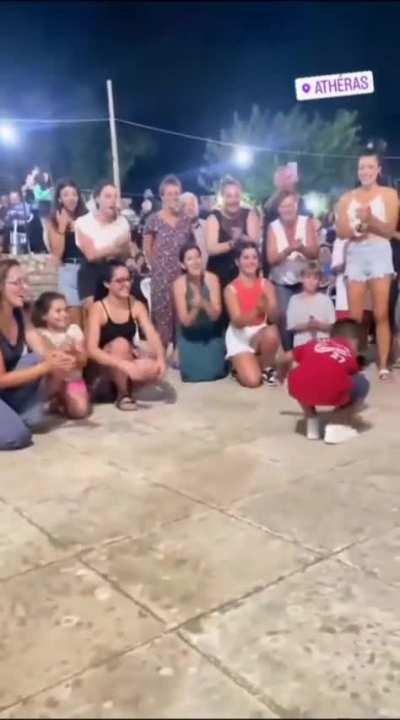 Kid performing a traditional Greek dance while friends and family cheer him on