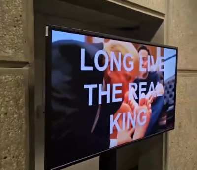 Monitors are hacked at the U.S. Department of Housing and Urban Development (HUD) to display an AI video of Trump licking Elon Musk’s toes. The caption reads: “LONG LIVE THE REAL KING.”