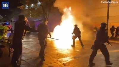 Liberal Antifa/Black Lives Matter/anarcho-leftist protestors assault police during the highly coordinated 100 Days Portland Riot. One protestor throws a Molotov cocktail at police; the mostly peaceful protestors cheer as it reaches its target.