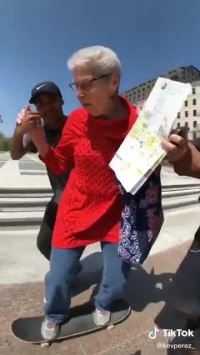 Two young skaters help an older woman skateboard for the first time and she's stoked