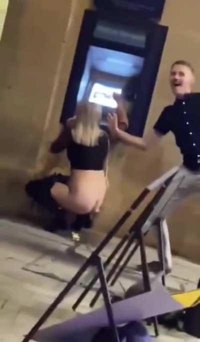 A woman publicly peeing on the street floor while using a ATM