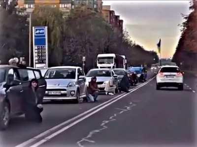 Ukrainians kneel on the road while in traffic as their fallen soldiers return home