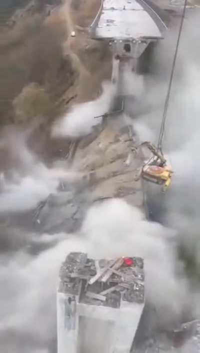 An excavator secured to a crane demolishing a bridge