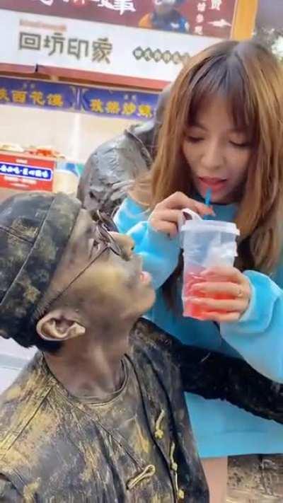 Woman gives drinks to a living statue street performer in a wierd way by using straw.