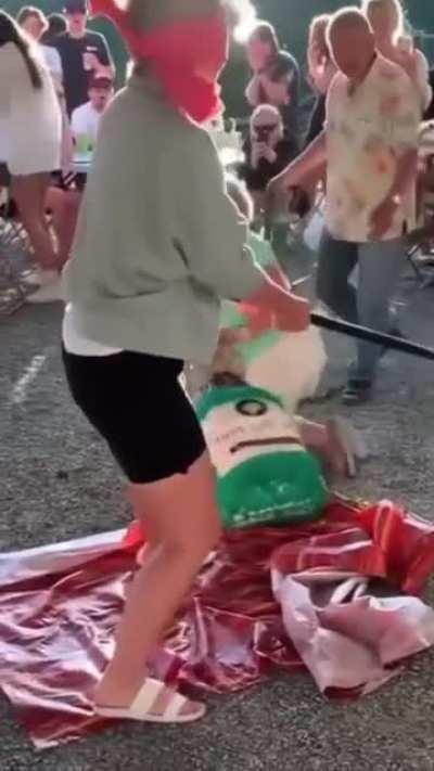 Oblivious wine aunt holds their hand out in front of a blindfolded partygoer to stop them from swinging at a pinata.