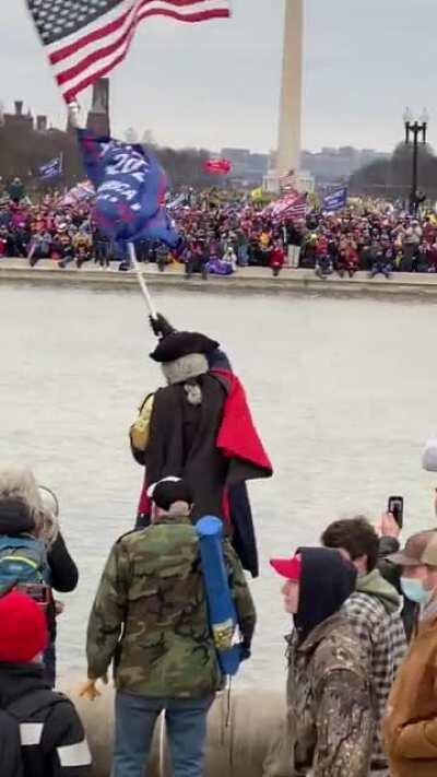 Man dressed in revolutionary garb carrying Trump's flag slogs through the water to call Trump a man of God. Cameraman quips: 