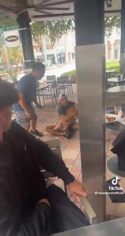 A large dog grabs a puppy off of a woman's lap while she is eating lunch