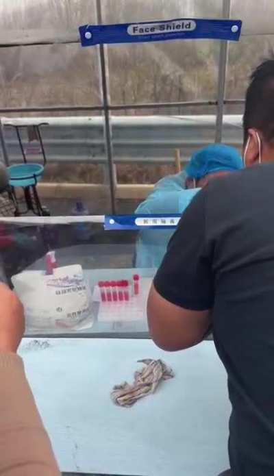 A nucleic acid test station worker casually smoking a cigarette, mask down, blowing smoke down people’s throats and sneezing😅🤣